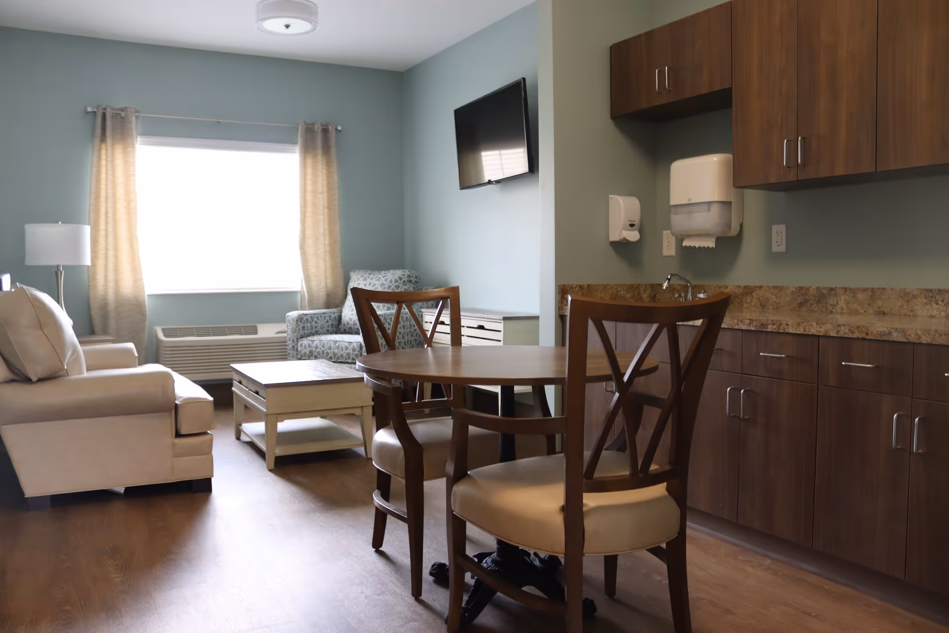 A cozy living area in an assisted living facility featuring a beige sofa, a patterned armchair, a wooden coffee table, a round wooden dining table with two chairs, a wall-mounted TV, and wooden cabinets with a countertop and a sink. The room has light blue walls, a window with beige curtains, and wood flooring.