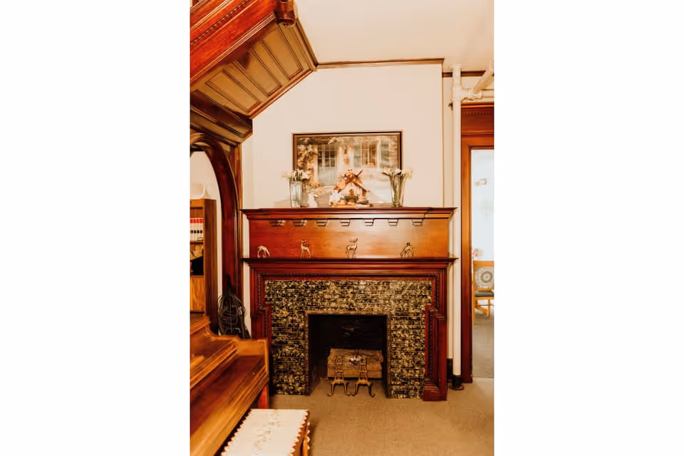 Ornate wooden fireplace mantel with decorative tile surround, vases, and framed artwork in a cozy interior room.