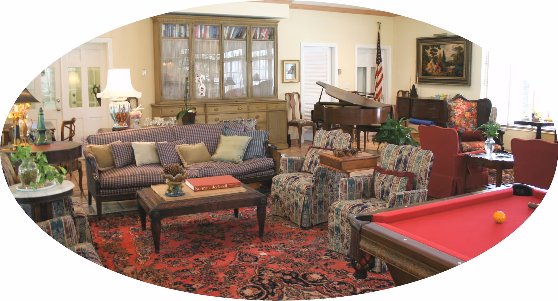 A cozy living room area with patterned sofas and armchairs, a red pool table, a wooden coffee table with books, a grand piano, various lamps, plants, and a large cabinet filled with books. The room has a warm and inviting atmosphere with a red patterned rug and an American flag in the corner.
