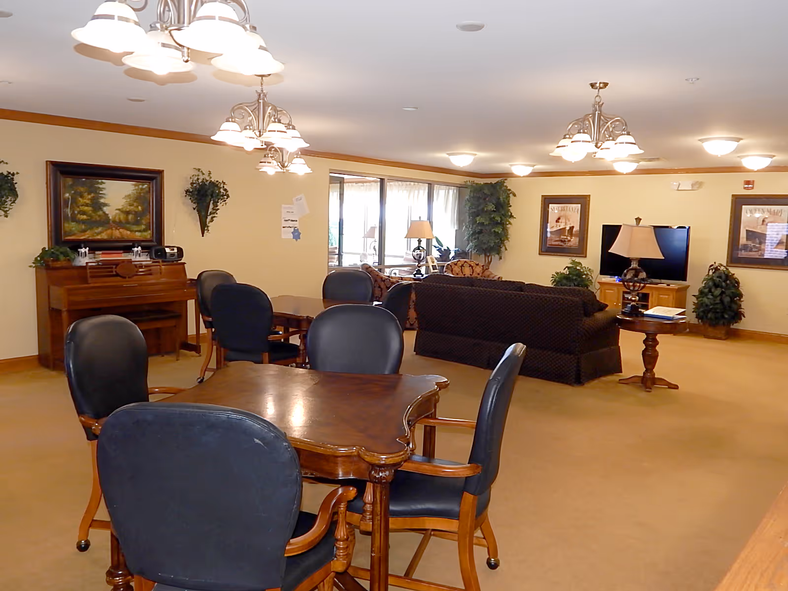 A spacious senior living common area with multiple tables and chairs in the foreground, a piano against the wall with a framed painting above it, and a seating area with couches, a TV, and lamps in the background. The room has beige walls, carpeted floor, and several ceiling light fixtures.