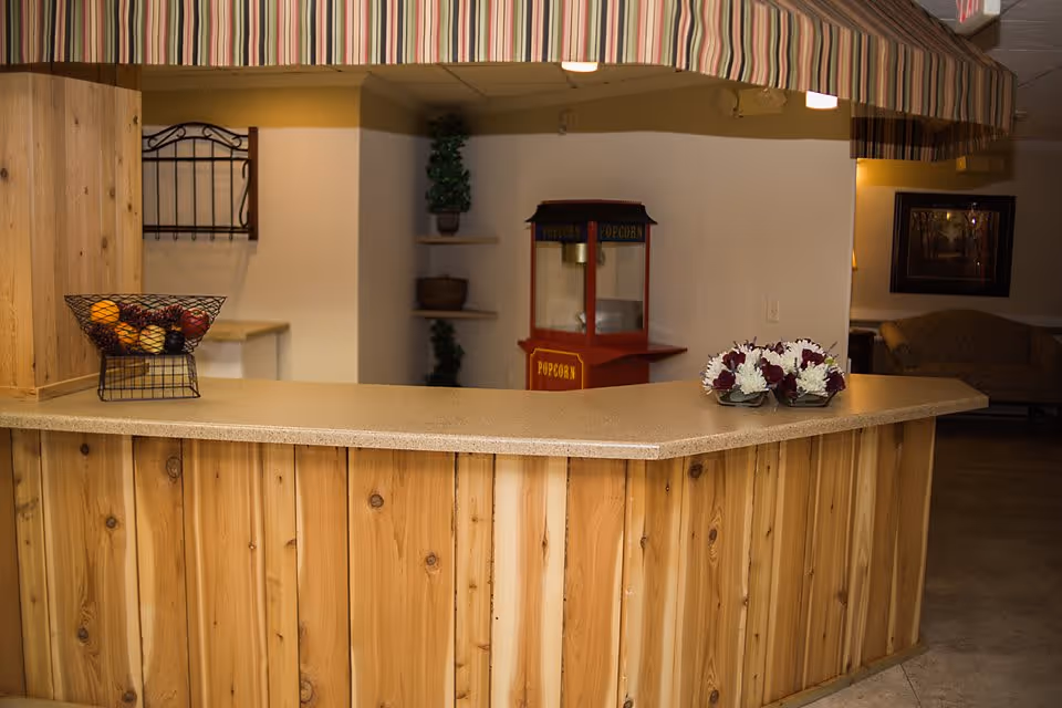 Interior view of a wooden reception or snack counter with a striped awning above. On the counter, there is a wire basket with artificial fruit and a small arrangement of white and purple flowers. In the background, there is a red popcorn machine and some shelves with potted plants. A couch and framed artwork are visible in a dimly lit adjacent room.