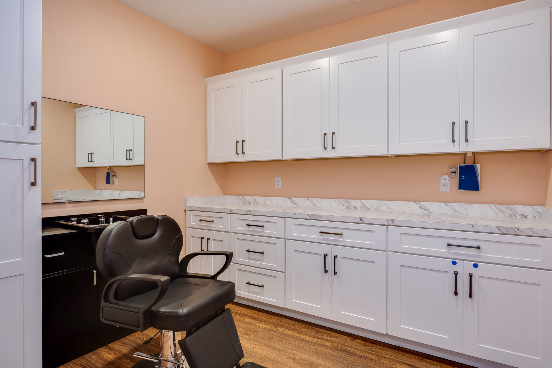 Interior view of a room with white cabinets and drawers along the wall, a marble countertop, a large mirror, and a black salon chair in front of a small black sink unit. The walls are painted peach, and the floor has wood-like flooring.