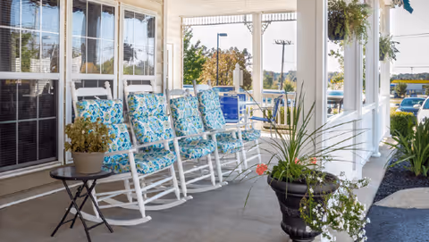 A covered outdoor porch area with six white rocking chairs lined up, each with blue and green patterned cushions. There are potted plants on a small black table and in a large black planter, with a view of trees, parked cars, and a street in the background.