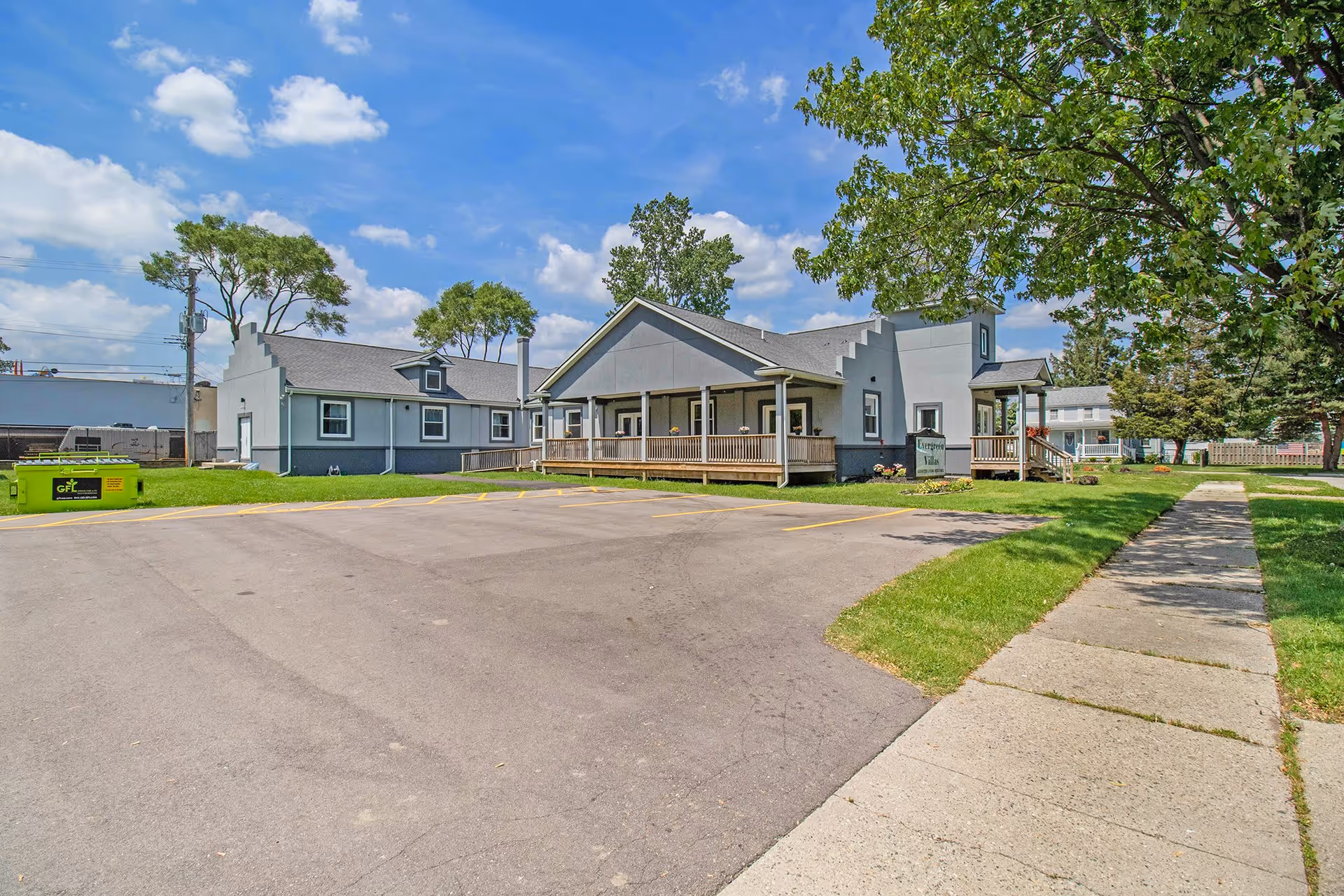 Exterior view of Evergreen Villas facility showing a gray building with a porch, surrounded by green grass and trees under a blue sky with some clouds. There is a paved parking lot and a sidewalk leading to the building.