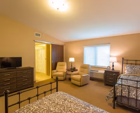 Well-lit assisted living bedroom with two beds, two recliners, a dresser with a TV, and a window with blinds.