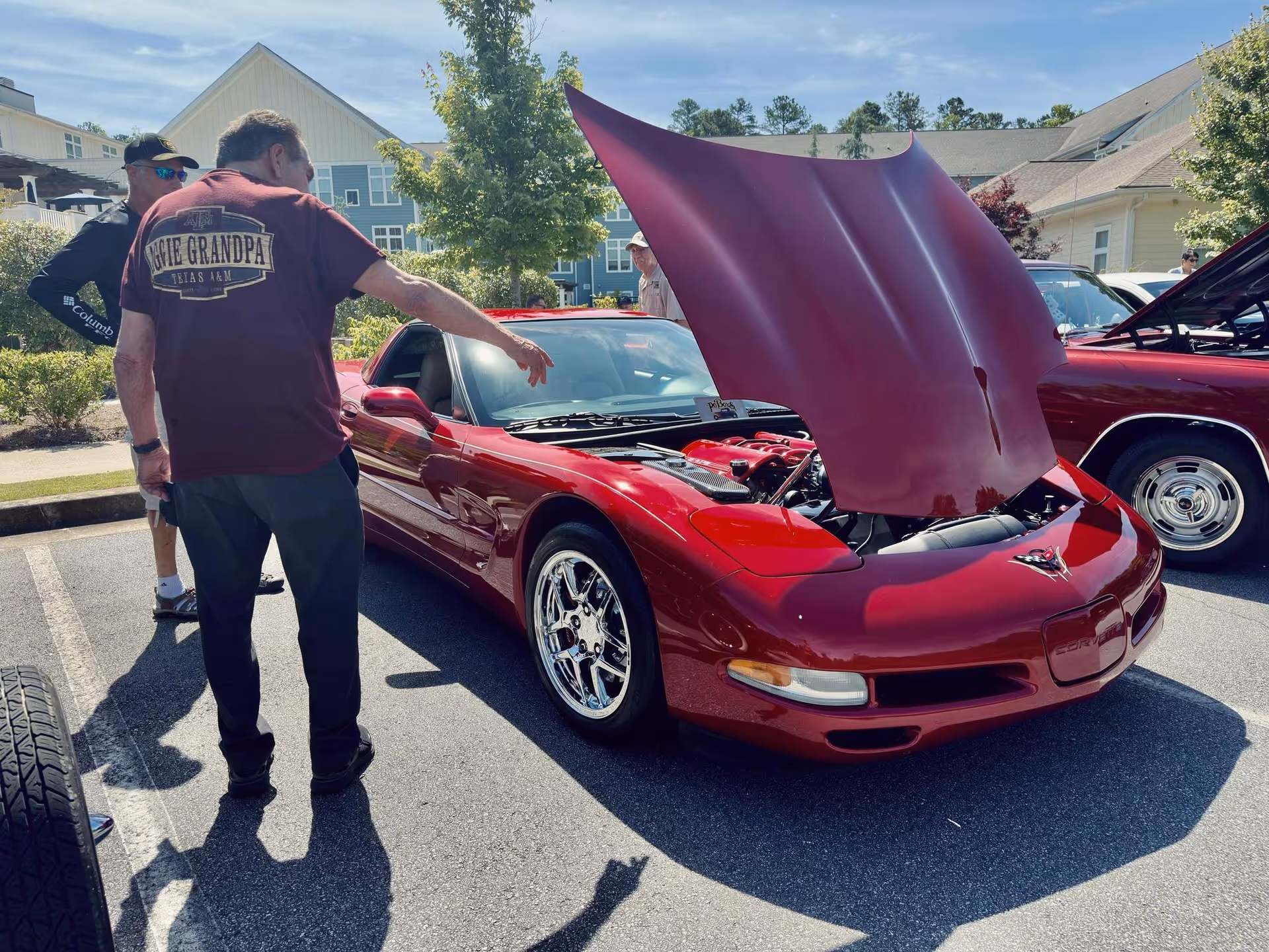 A man wearing a maroon shirt with the text 'Aggie Grandpa Texas A&M' is looking at a red sports car with its hood open in a parking lot. Another man stands nearby. Residential buildings and trees are visible in the background under a clear sky.