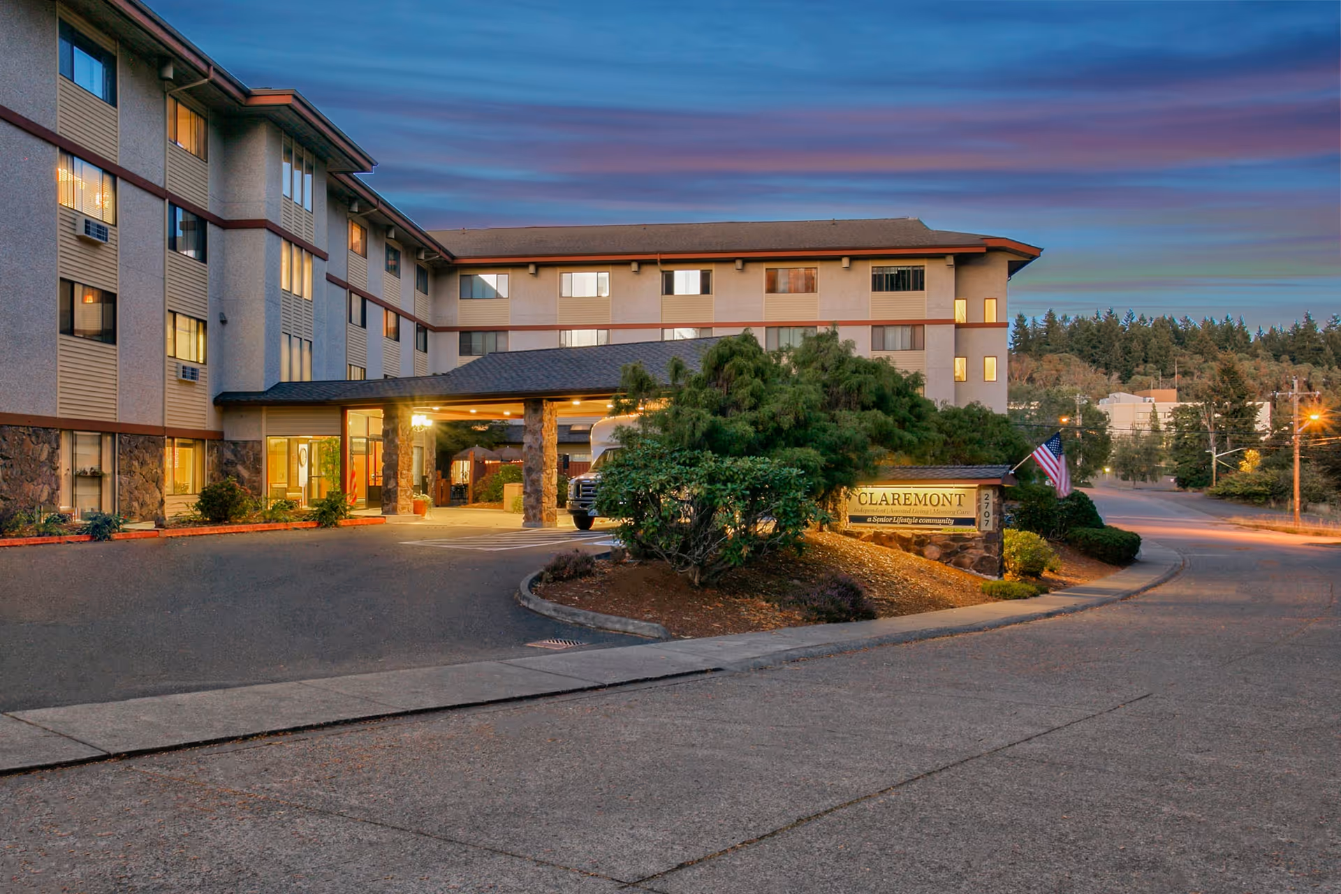 Exterior view of a senior living facility building at dusk with a covered entrance supported by stone pillars, surrounded by greenery and a sign that reads 'CLAREMONT Independent | Assisted Living | Memory Care a Senior Lifestyle community'. An American flag is displayed near the sign.