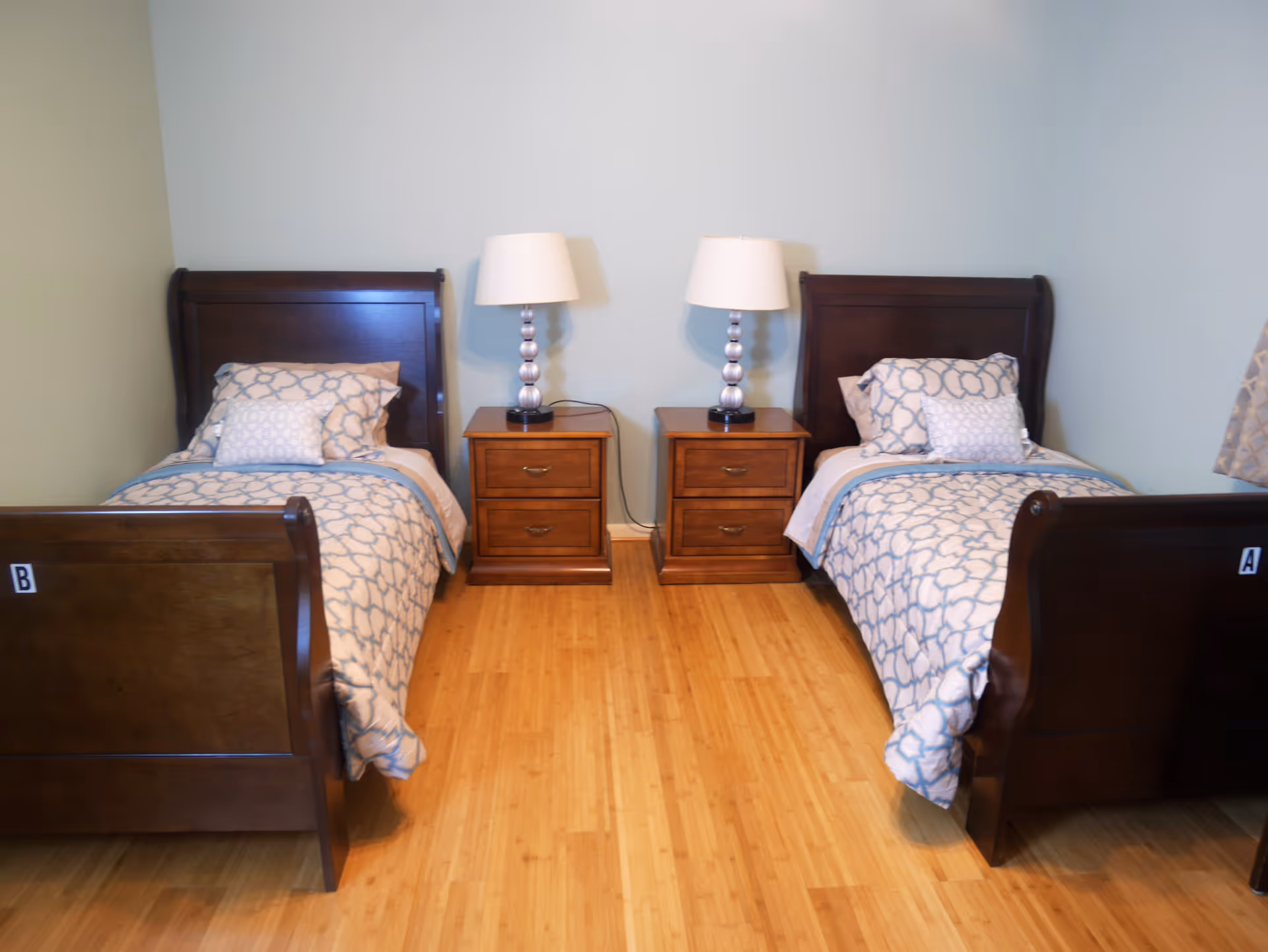 A bedroom with two twin beds, each with a dark wooden headboard and footboard. Both beds have matching blue and white patterned bedding and pillows. Between the beds are two wooden nightstands, each with a lamp featuring a silver base and white shade. The room has light blue walls and a wooden floor.