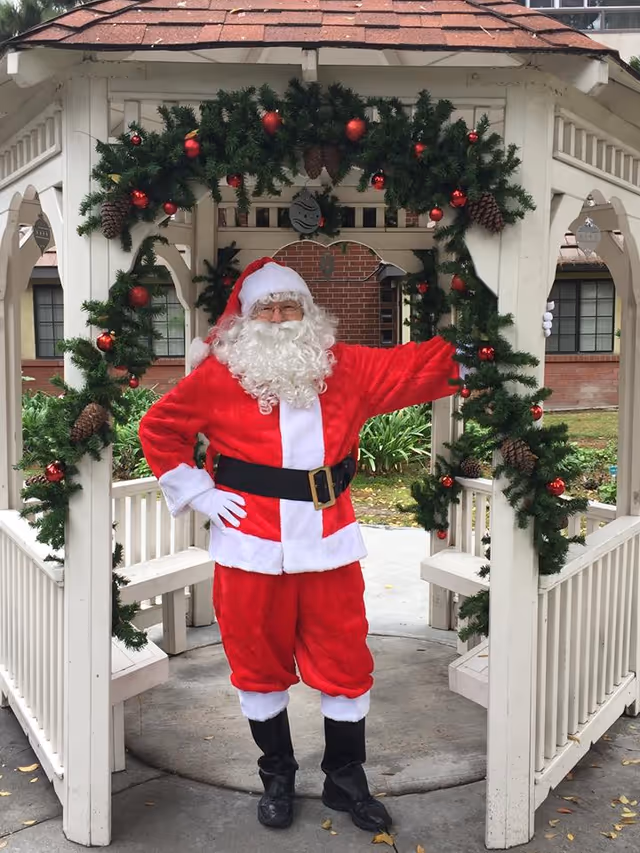 A person dressed as Santa Claus stands inside a white gazebo decorated with green garlands, pine cones, and red ornaments. The background shows a brick building and some greenery.