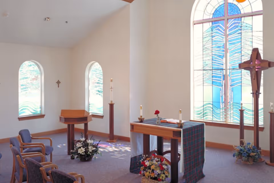 Interior of a small chapel with stained glass windows, a wooden altar covered with a cloth, a wooden lectern, several chairs, a wooden cross, candles, and floral arrangements.