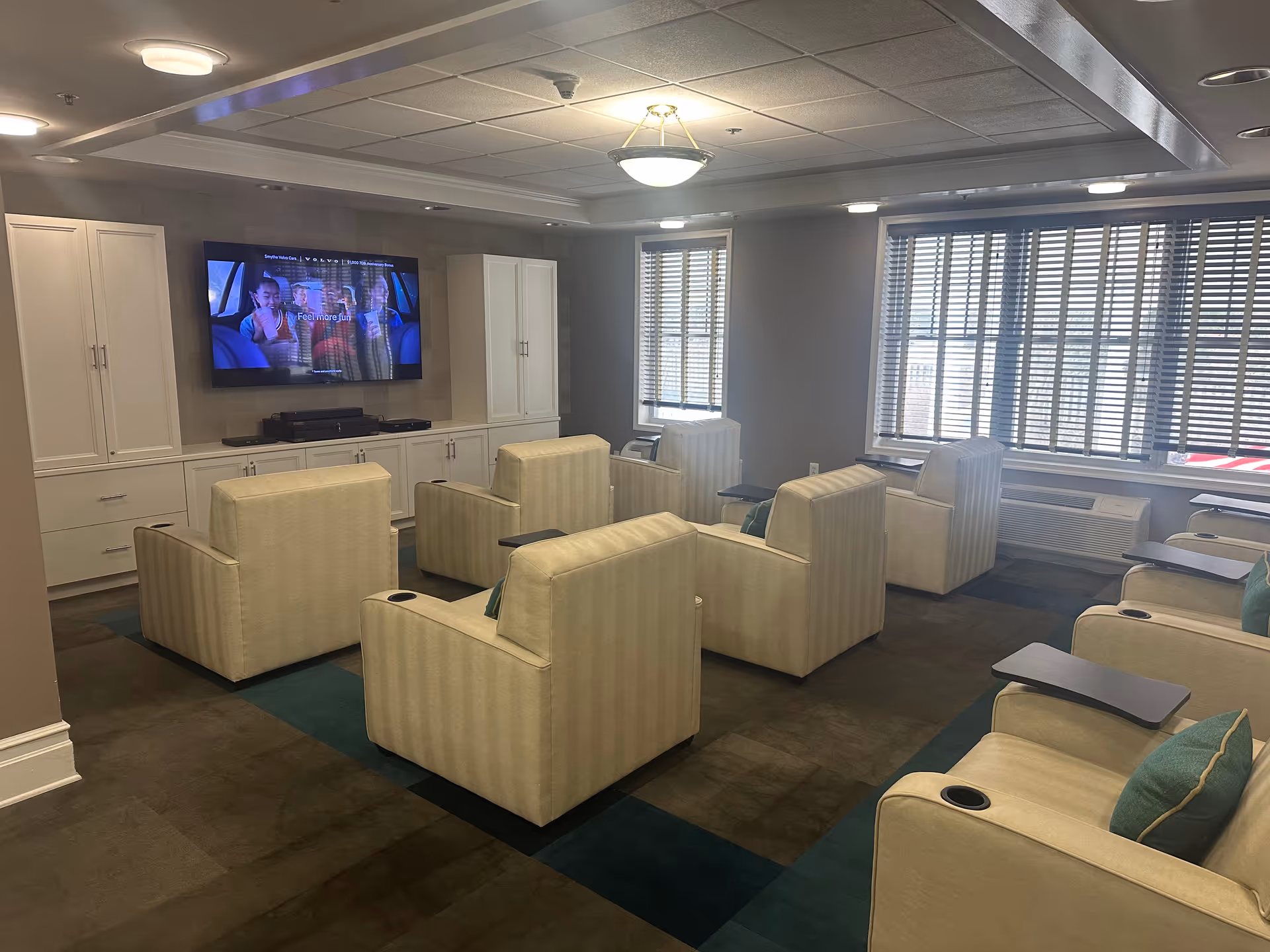 A cozy common area with multiple beige armchairs arranged in rows facing a large flat-screen TV mounted on a wall. The room has large windows with blinds, a carpeted floor, and built-in white cabinetry beneath the TV. The ceiling features recessed lighting and a central light fixture.