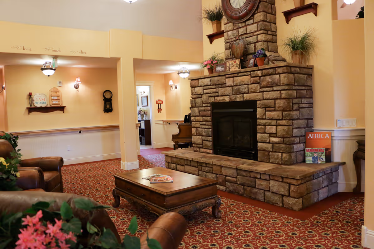 A cozy living room area in a senior living facility featuring a large stone fireplace with decorative plants and framed pictures on the mantel. There are leather armchairs and a wooden coffee table with magazines on it. The room has warm yellow walls, patterned red carpet, and soft lighting from wall sconces and ceiling fixtures. A piano is visible in the background along with a hallway leading to other rooms.