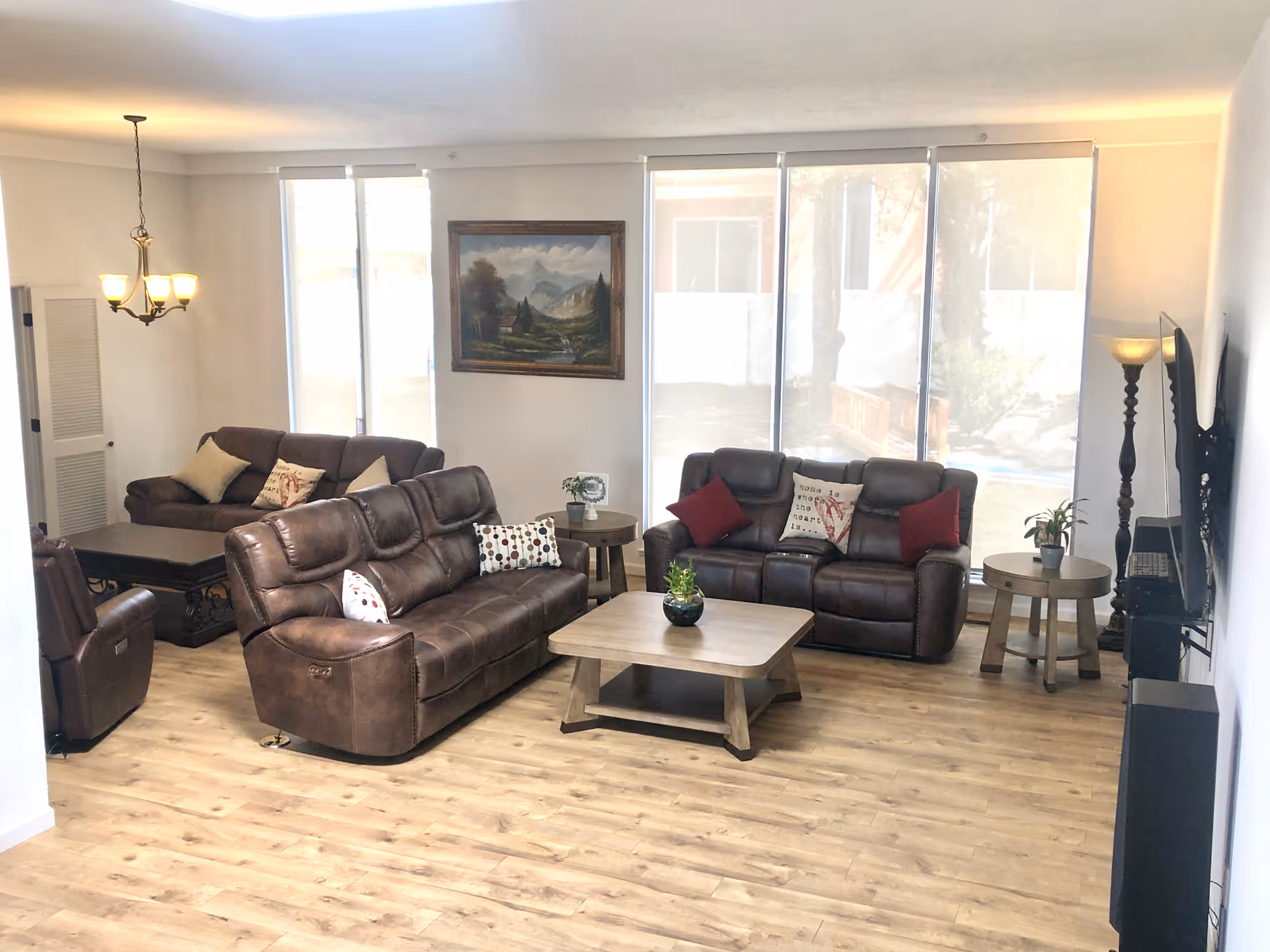 A bright living room with large windows covered by sheer blinds, featuring two brown leather sofas and a matching armchair. There are decorative pillows on the sofas, a wooden coffee table with a small plant, side tables with plants and a lamp, a wall-mounted TV, and a framed landscape painting on the wall.
