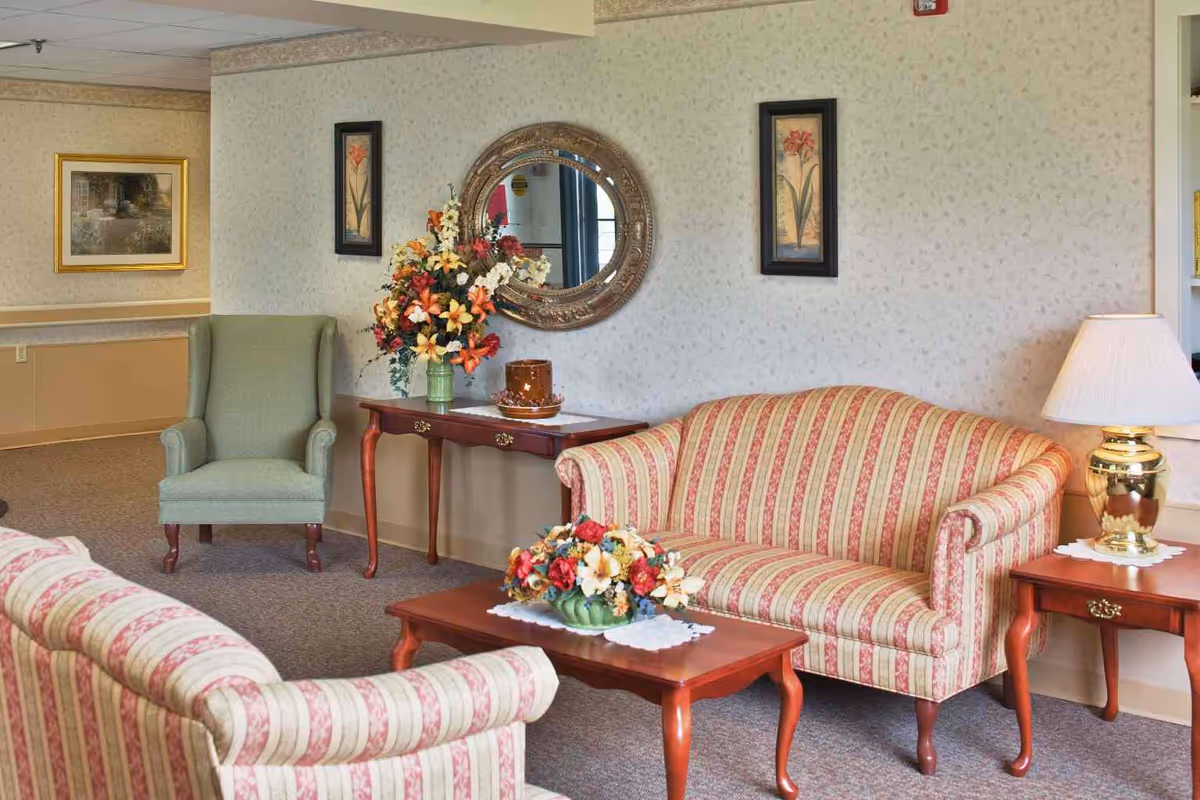 A cozy living room area with two striped sofas facing each other, a green armchair, a wooden coffee table with a floral arrangement, a side table with a gold lamp, and a console table with a vase of flowers and a decorative item. The walls are decorated with framed floral artwork and a round mirror.