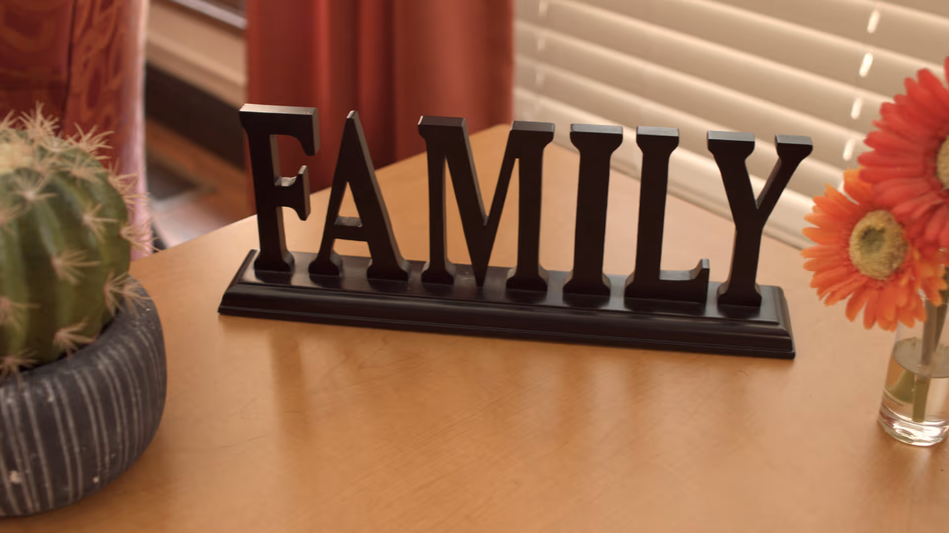A wooden table with a black decorative sign spelling the word 'FAMILY' in capital letters. To the left of the sign is a small potted cactus, and to the right is a small glass vase holding orange and red flowers. In the background, there are window blinds and a red curtain.