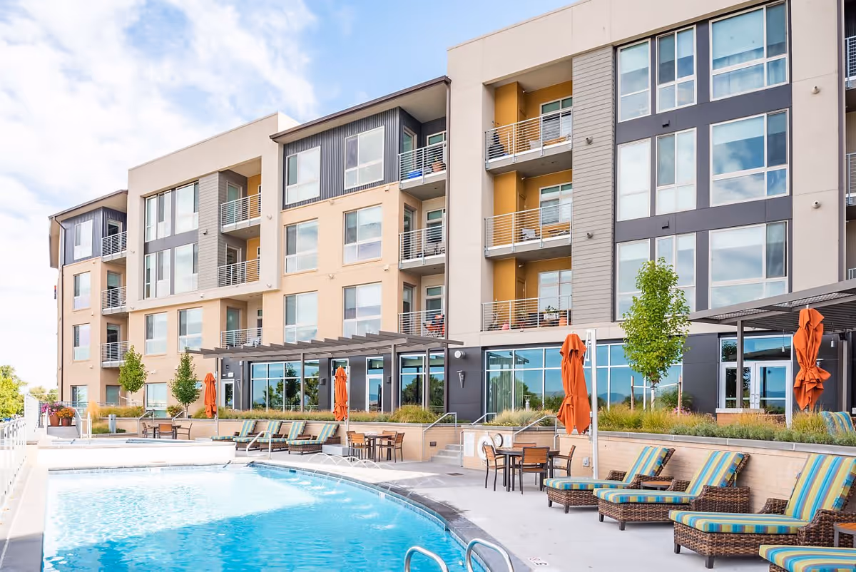 Outdoor swimming pool area with lounge chairs and tables with umbrellas in front of a multi-story residential building with balconies and large windows under a partly cloudy sky.