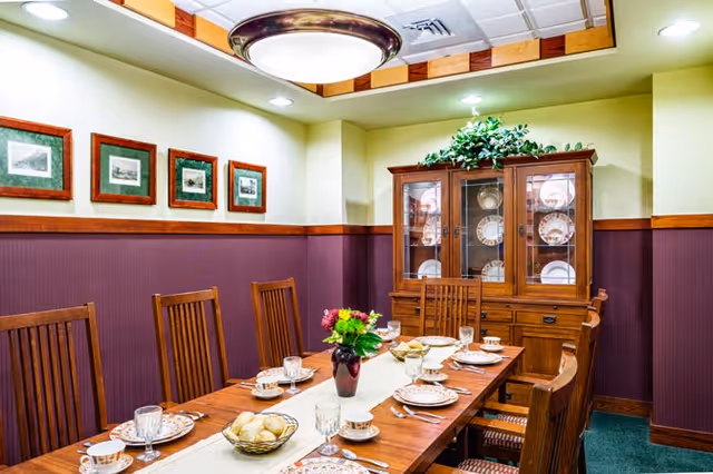 Dining room with a long wooden table set with plates, cups and glasses, wooden chairs, framed pictures on the wall and a china cabinet.
