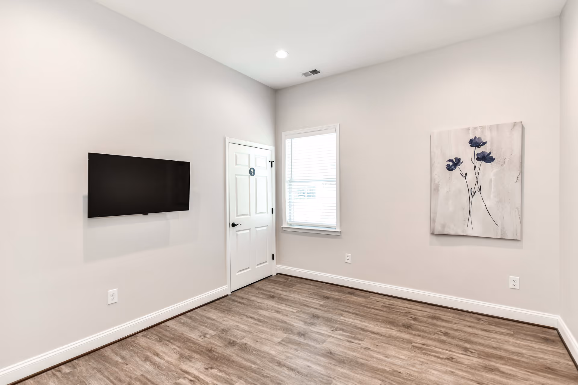 A small, empty room with light gray walls and wood flooring. A flat-screen TV is mounted on the left wall, a white door with a restroom sign is in the corner, and a window with blinds is next to the door. On the right wall, there is a painting of three blue flowers on a light background. The room is well-lit with a ceiling light.