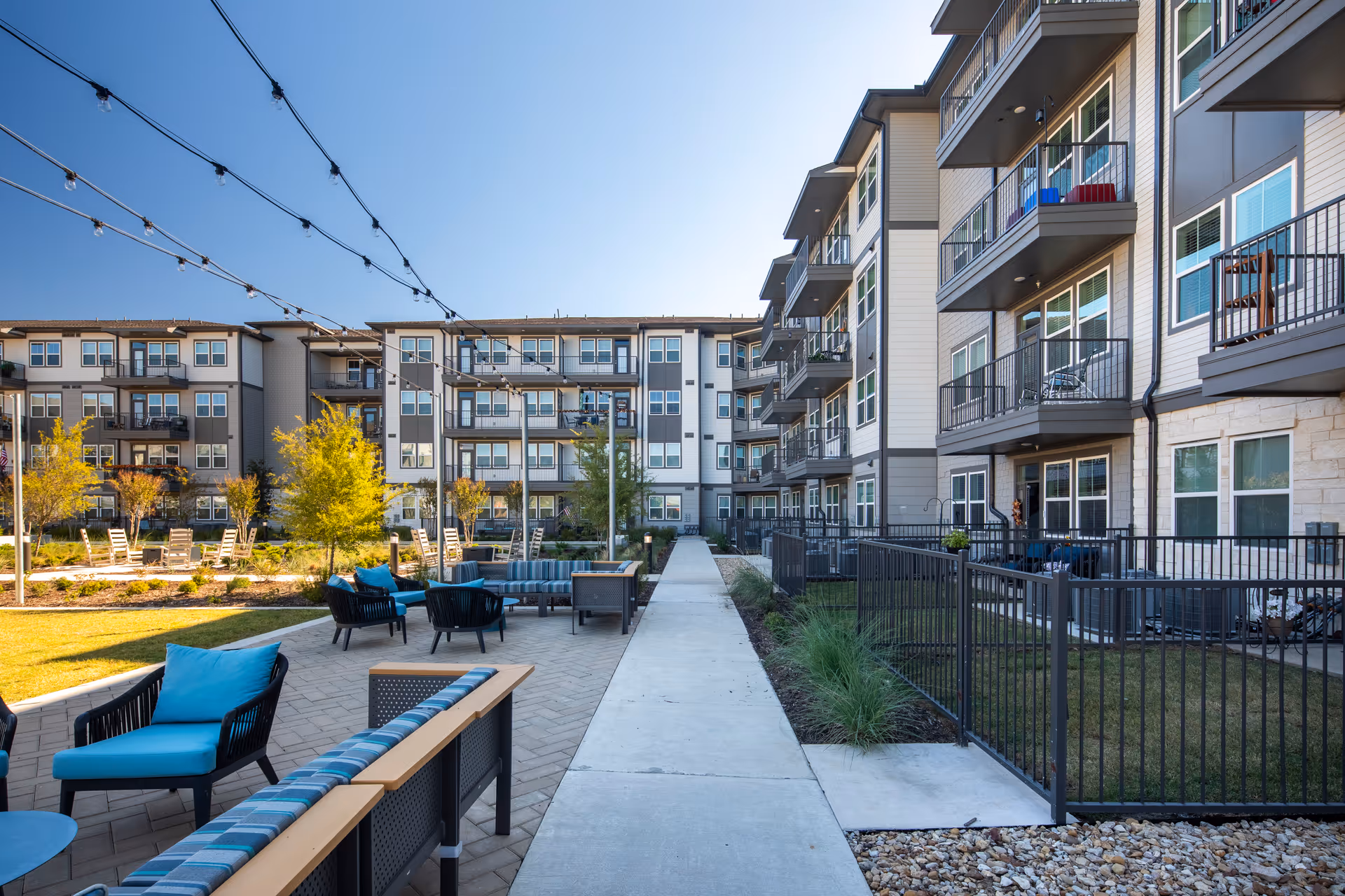 Outdoor courtyard area of a senior living facility with modern multi-story buildings surrounding it. The courtyard features a paved walkway, seating areas with blue cushioned chairs and sofas, string lights overhead, small trees, and landscaped greenery under a clear blue sky.