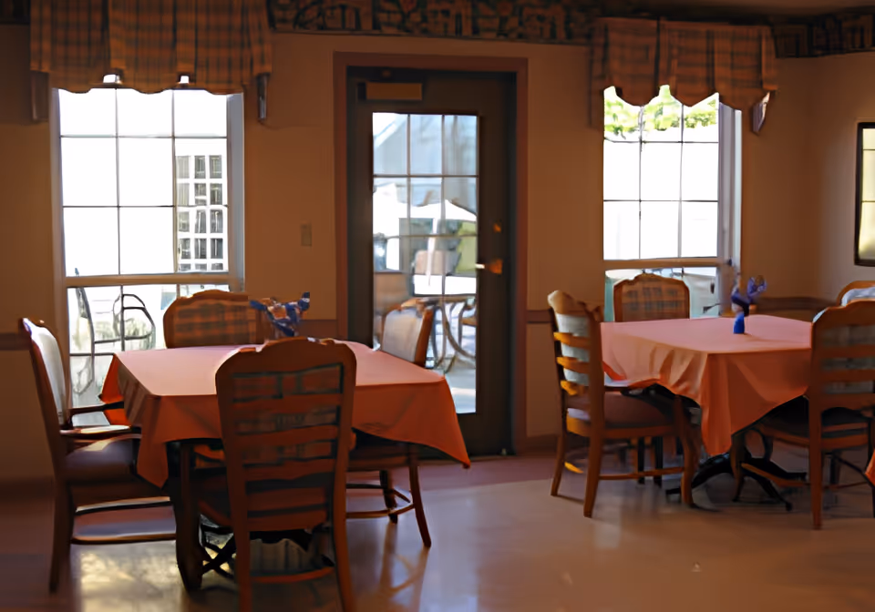 Interior view of a dining room with two tables covered with orange tablecloths. Each table is surrounded by wooden chairs with cushioned seats and backs. The room has large windows with checkered valances and a glass door leading outside. Small decorative items are placed on the tables.