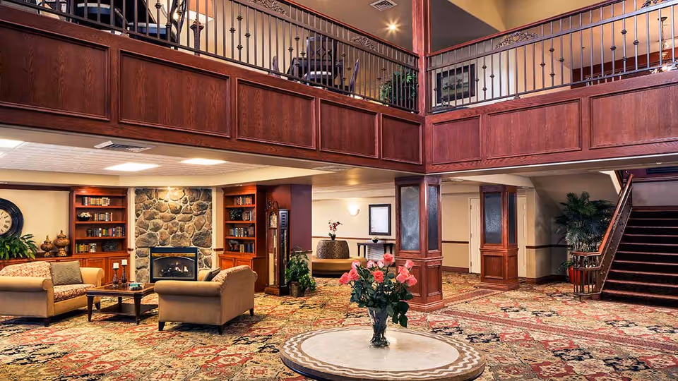 Spacious senior living facility common area with a patterned carpet, two beige sofas around a wooden coffee table, a stone fireplace flanked by built-in bookshelves, a round table with a vase of pink flowers in the foreground, and a staircase leading to an upper level with a wooden railing.