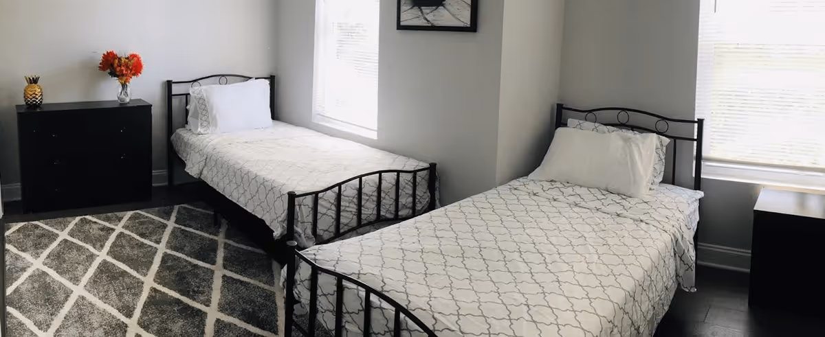 A bedroom with two single beds featuring black metal frames and white patterned bedding. Each bed has a white pillow. Between the beds is a small black nightstand, and to the left is a black dresser with a vase of red flowers and a decorative pineapple on top. The room has two windows with white blinds and a gray and white patterned rug on the floor.