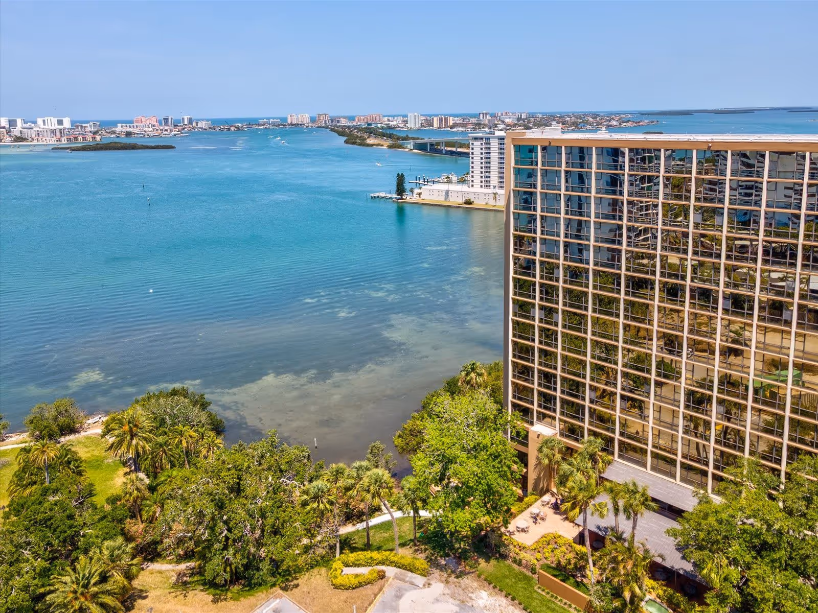 Aerial view of a waterfront area with a large building featuring reflective glass windows on the right side. The building is surrounded by lush green trees and palm trees. In the background, there is a wide body of blue water with boats and a distant city skyline under a clear blue sky.