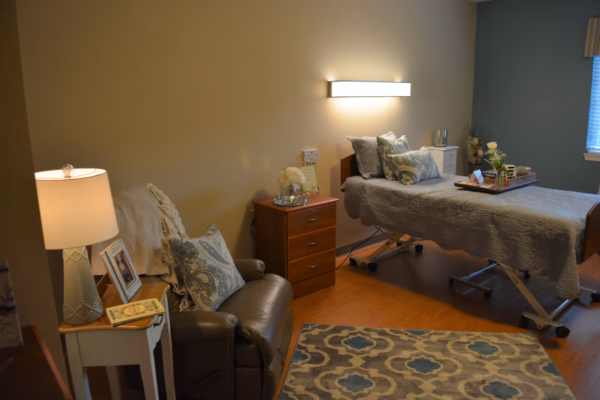 A cozy bedroom in a senior living facility with a single bed covered in a gray quilt and multiple patterned pillows. A wooden nightstand with a flower vase and a framed note is beside the bed. There is a comfortable leather armchair with a decorative pillow and a blanket draped over it, next to a small white side table with a lamp and a book. The room has wooden flooring and a patterned area rug, with a window partially covered by blinds on the right side.