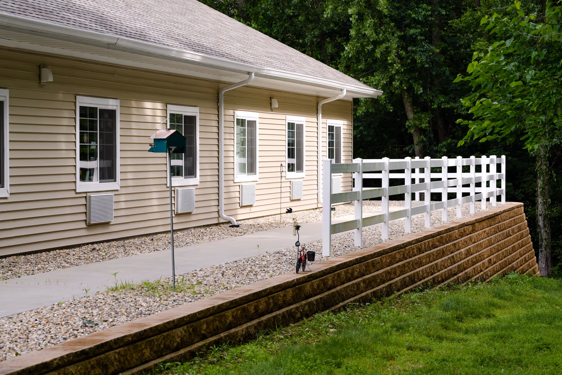 Side exterior of a beige single-story building with windows, a white fence along a raised retaining wall, a paved walkway and surrounding trees.
