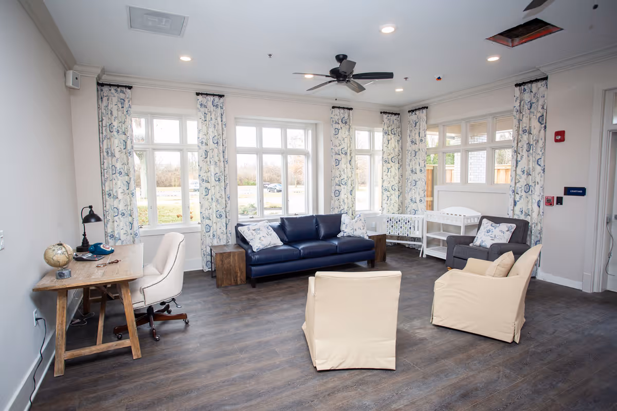 A bright living room with large windows covered by floral curtains. The room features a dark blue sofa with patterned pillows, a gray armchair, two beige chairs, and a wooden desk with a white office chair. There is a ceiling fan and wooden flooring, with a changing table and baby crib near the windows.