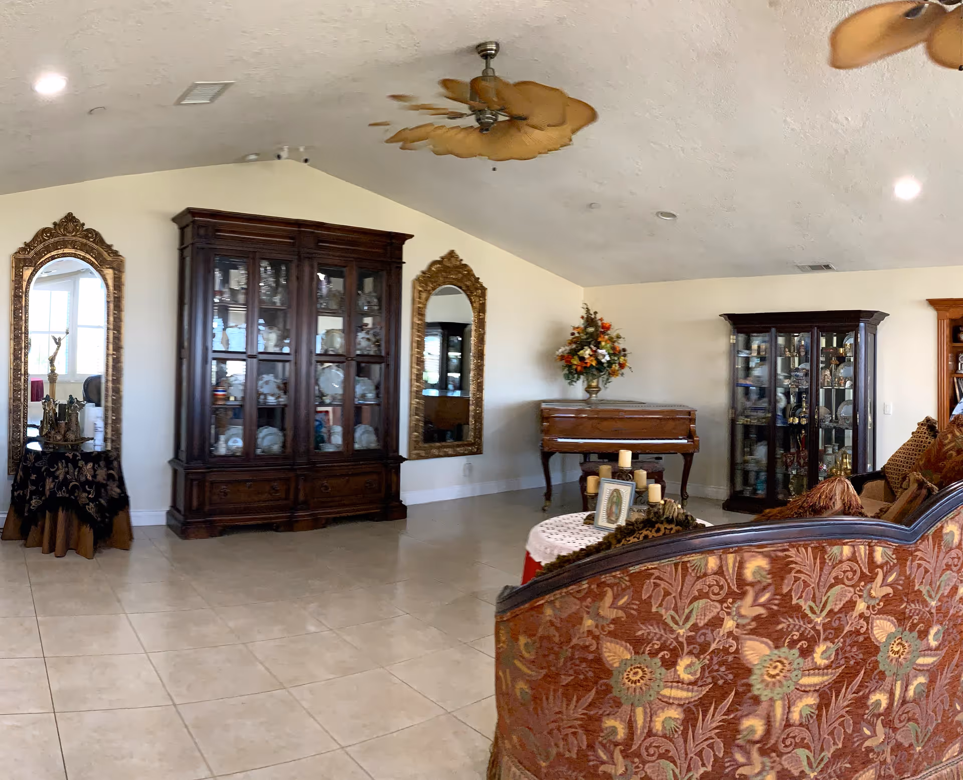 Spacious living room with tiled floor, an ornate sofa in the foreground, glass display cabinets, large gilded mirrors, and a piano against the wall.