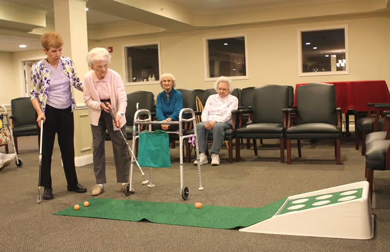 Two elderly women playing indoor mini golf on a green putting mat with a ramp in a common room. Two other elderly women are seated in chairs watching the game. The room has beige walls, carpeted floor, and several chairs arranged along the walls.