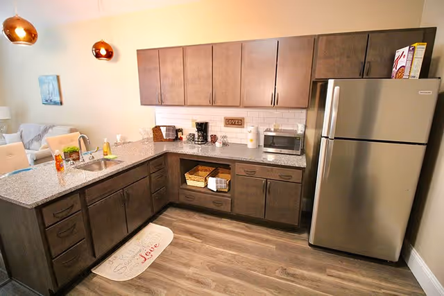 A modern kitchen area with dark wood cabinets, a stainless steel refrigerator, a granite countertop with a sink, a coffee maker, a microwave, and decorative items including a small sign that says 'LOVED'. The kitchen opens to a living space with a sofa and chairs visible in the background.