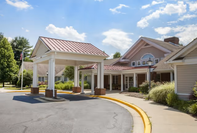 Exterior view of a senior living facility entrance with a covered drop-off area supported by white columns with brick bases. The building has beige siding, a red metal roof, and landscaped greenery around the entrance. The sky is blue with scattered clouds.