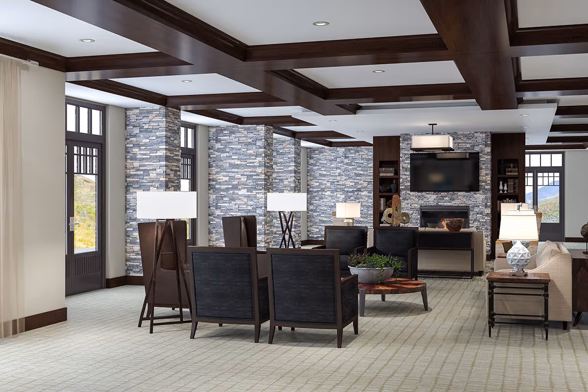 A spacious and modern living room area with stone accent walls, large windows, and dark wood ceiling beams. The room features multiple seating arrangements including dark wood chairs with black cushions, beige sofas, and several floor and table lamps. A flat-screen TV is mounted above a fireplace on the far wall, with decorative items on shelves and tables. The room has a light-colored carpet and large windows letting in natural light.
