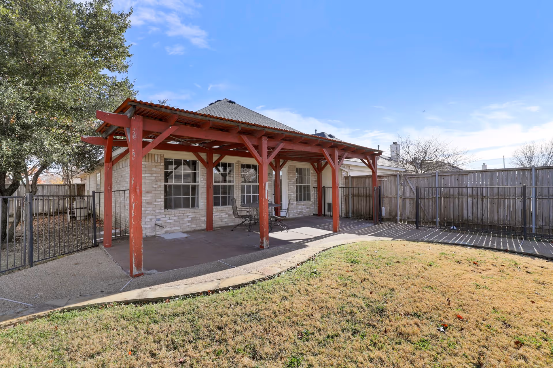 Outdoor patio area with a red wooden pergola attached to a brick building. There are two metal chairs and a small table under the pergola. The patio is surrounded by a metal fence and a wooden privacy fence, with a grassy lawn in the foreground and trees on the left side.