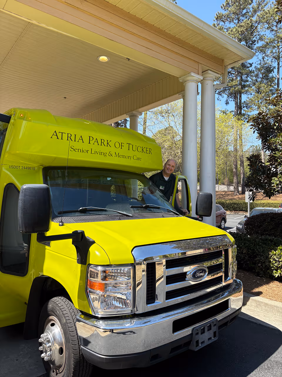 A bright yellow shuttle bus with the text 'Atria Park of Tucker Senior Living & Memory Care' parked under a covered entrance with white columns. A man is standing at the open driver's side door smiling. Trees and parked cars are visible in the background.