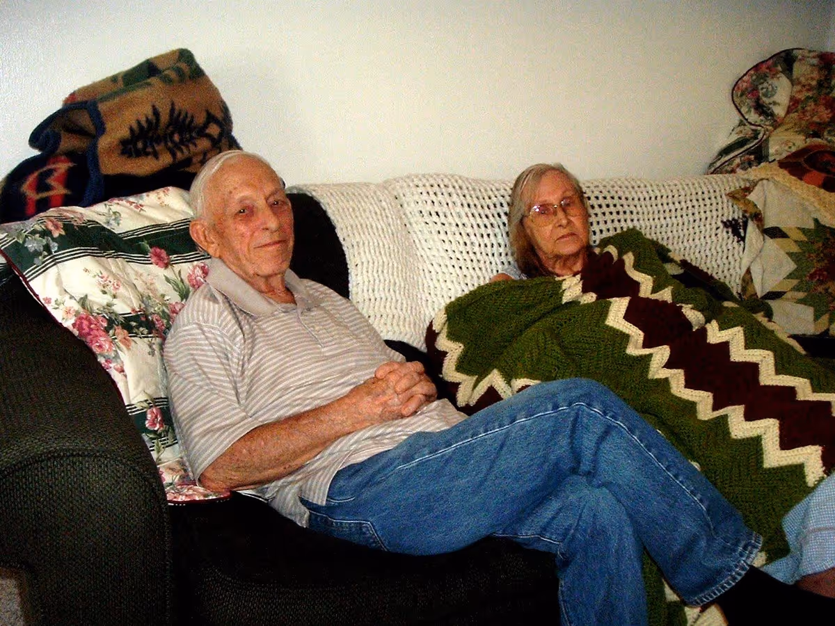 An elderly man and woman sitting on a dark-colored couch with floral and patterned pillows. The man is wearing a striped polo shirt and jeans, sitting with his hands clasped and smiling slightly. The woman is covered with a green, white, and maroon zigzag patterned blanket and wearing glasses, looking downwards. The background is a plain white wall.