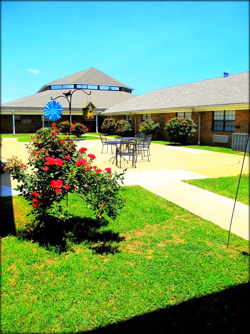 Outdoor courtyard area of a senior living facility with green grass, flowering bushes, a birdhouse, a blue decorative flower, and metal patio tables and chairs. The building surrounds the courtyard with brick walls and windows under a clear blue sky.