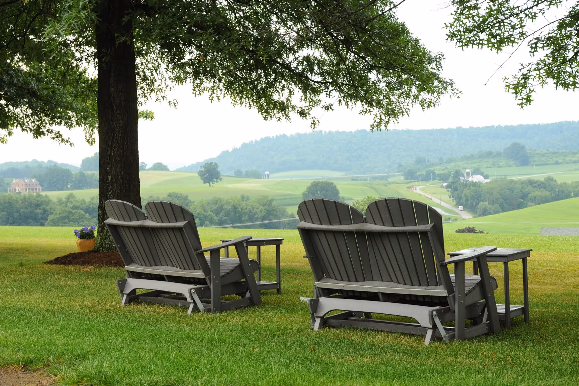 Two wooden benches with armrests and a small table between them are placed on a grassy lawn under a large tree. In the background, rolling green hills and a winding road are visible under a cloudy sky.