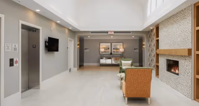 Well-lit lobby with elevators on the left, seating in the center, and a fireplace on the right beneath a skylight ceiling.
