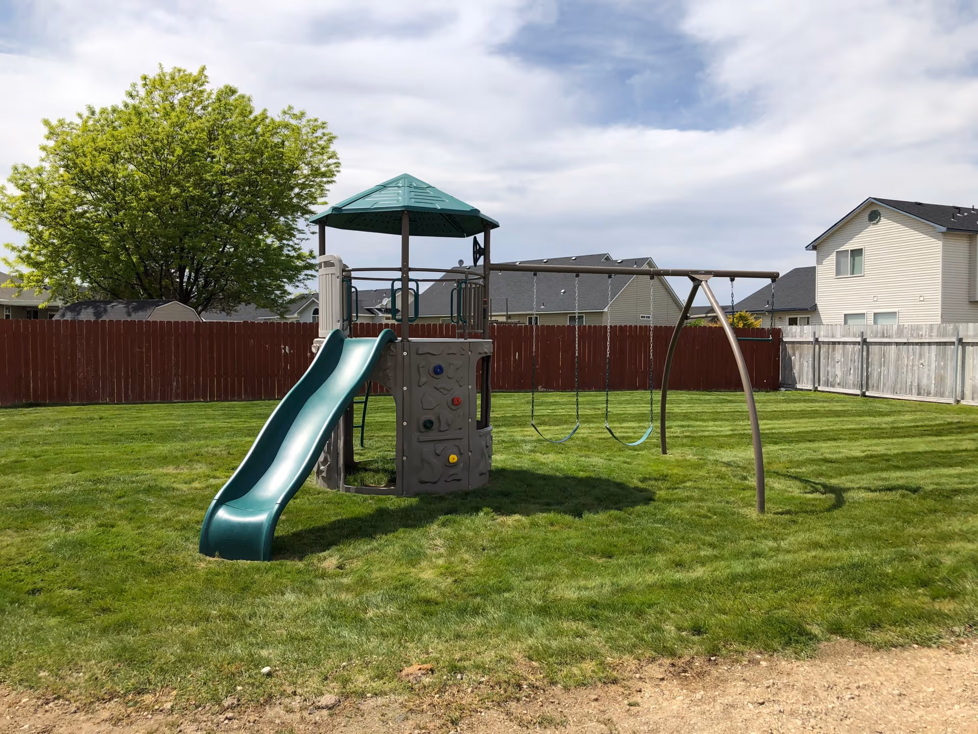 A playground set in a grassy fenced backyard with a slide, climbing wall, and two swings under a partly cloudy sky. Surrounding houses and a large green tree are visible in the background.