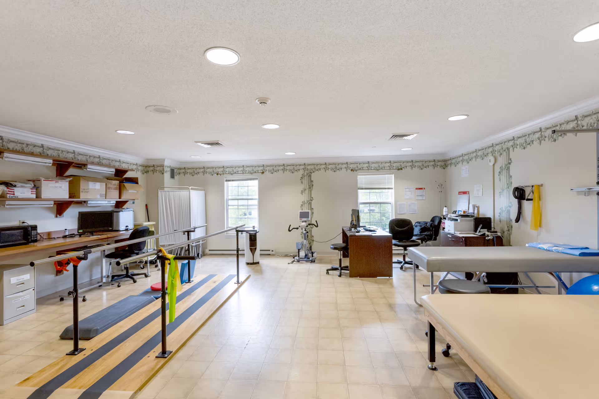A spacious rehabilitation or physical therapy room with parallel bars for walking exercises, therapy tables, exercise equipment, desks with chairs, and shelves with supplies. The room has two windows letting in natural light and light-colored walls with decorative trim near the ceiling.