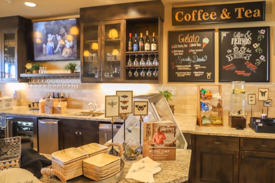 A cozy coffee and tea station in a senior living community with wooden cabinets, a granite countertop, and a display of wine bottles and glasses. There are chalkboard menus on the wall listing gelato flavors and specialty drinks. On the counter, there are stacks of wooden plates, small framed insect illustrations, a brochure titled 'Royal Chef,' and a water dispenser with lemon slices.