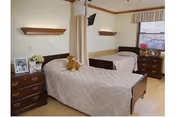 A shared bedroom in a nursing center with two single beds separated by a privacy curtain. Each bed has a wooden headboard and is covered with a beige quilt. There are two wooden dressers, one beside each bed, with personal items including framed photos, flowers, and a teddy bear on one bed. A window with blinds and a valance is visible in the background.
