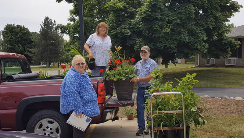 Three people outdoors near a red pickup truck with the tailgate down, holding and arranging potted plants and flowers. They are standing on a paved area with grass, trees, and buildings in the background.