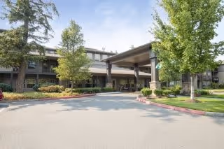Exterior view of a senior living facility with a covered entrance, surrounded by trees and landscaped greenery under a clear sky.