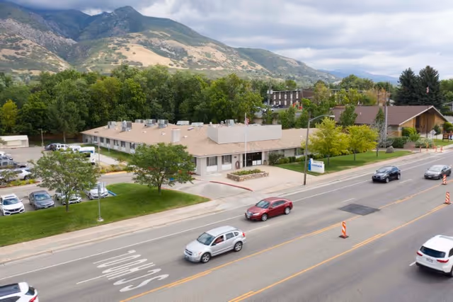 Aerial view of a single-story healthcare building front next to a road with cars, landscaped lawn and mountains in the background.