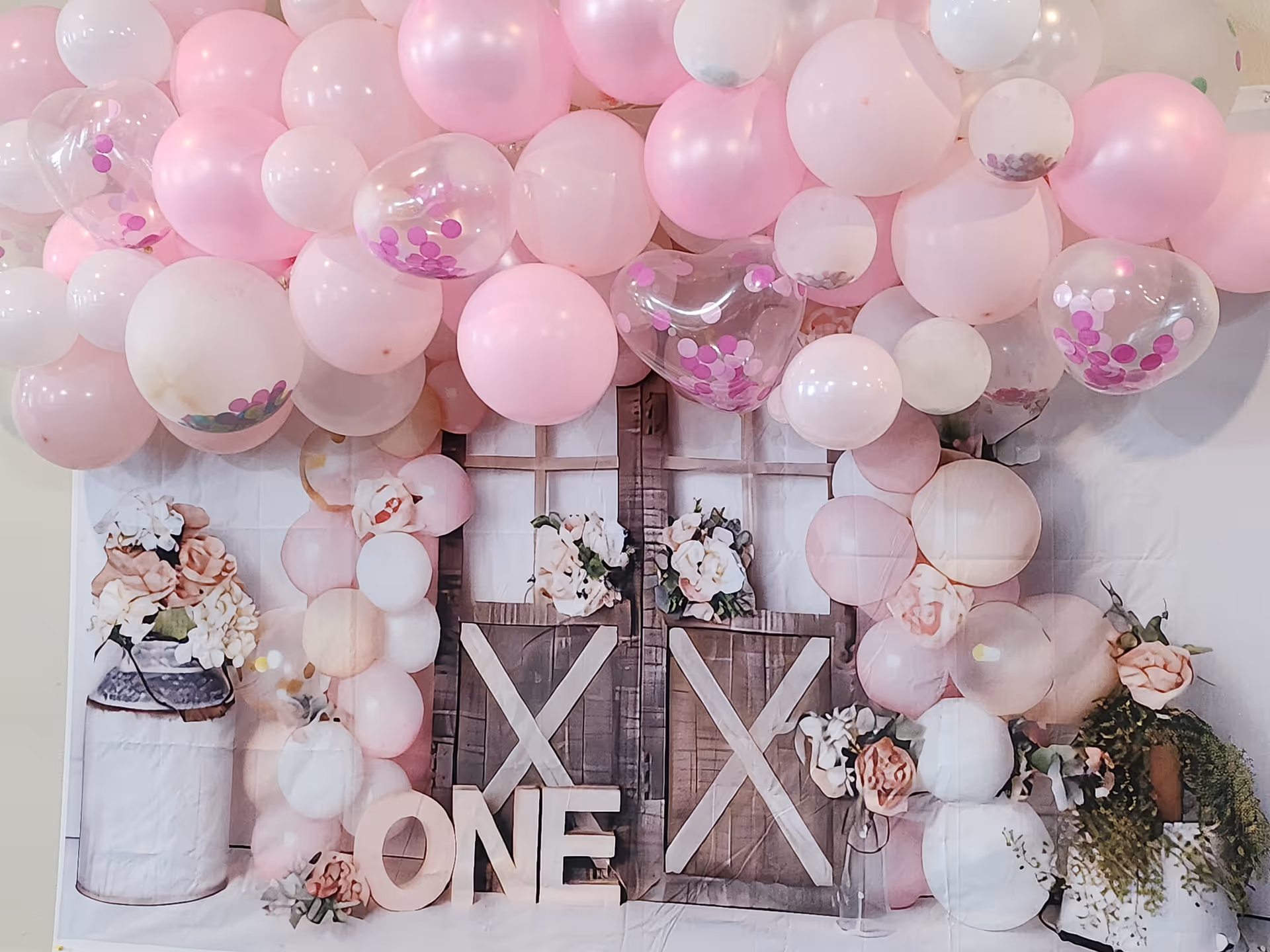 Decorative setup with pink, white, and transparent balloons with pink confetti, arranged around a rustic wooden window frame backdrop adorned with flowers and the word 'ONE' in large wooden letters, suggesting a first birthday or anniversary celebration.