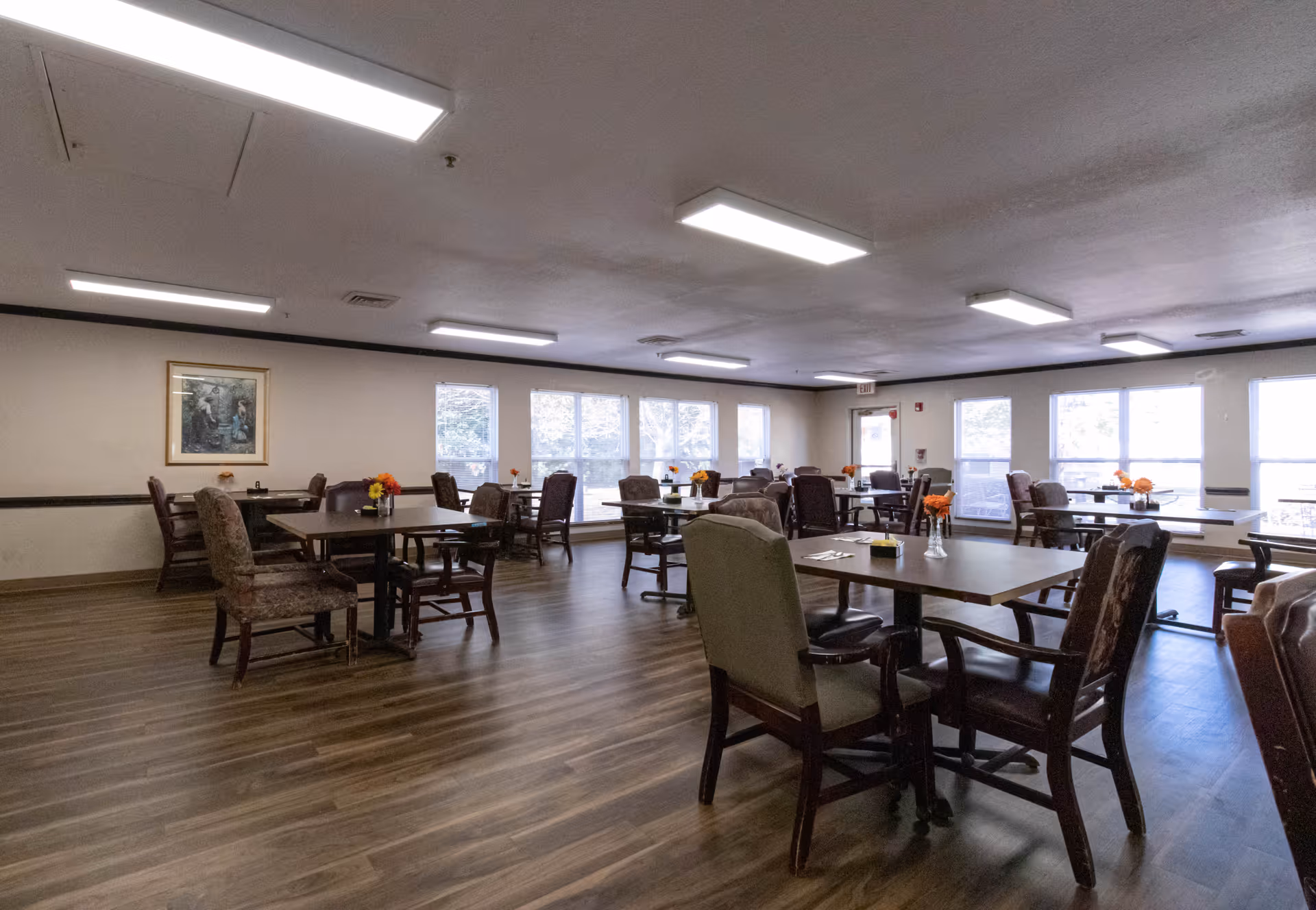 A spacious dining room with multiple tables and chairs arranged neatly. Each table has a small vase with orange flowers. Large windows along one wall allow natural light to fill the room. The floor is wood, and the ceiling has rectangular fluorescent lights. A framed picture hangs on one wall.